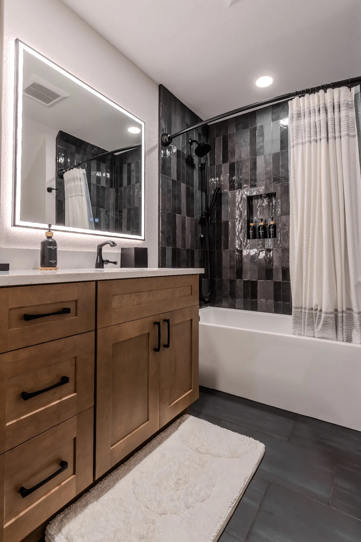 Remodeled hall bathroom with charcoal zellige tile tub surround, stained maple vanity, and backlit LED mirror