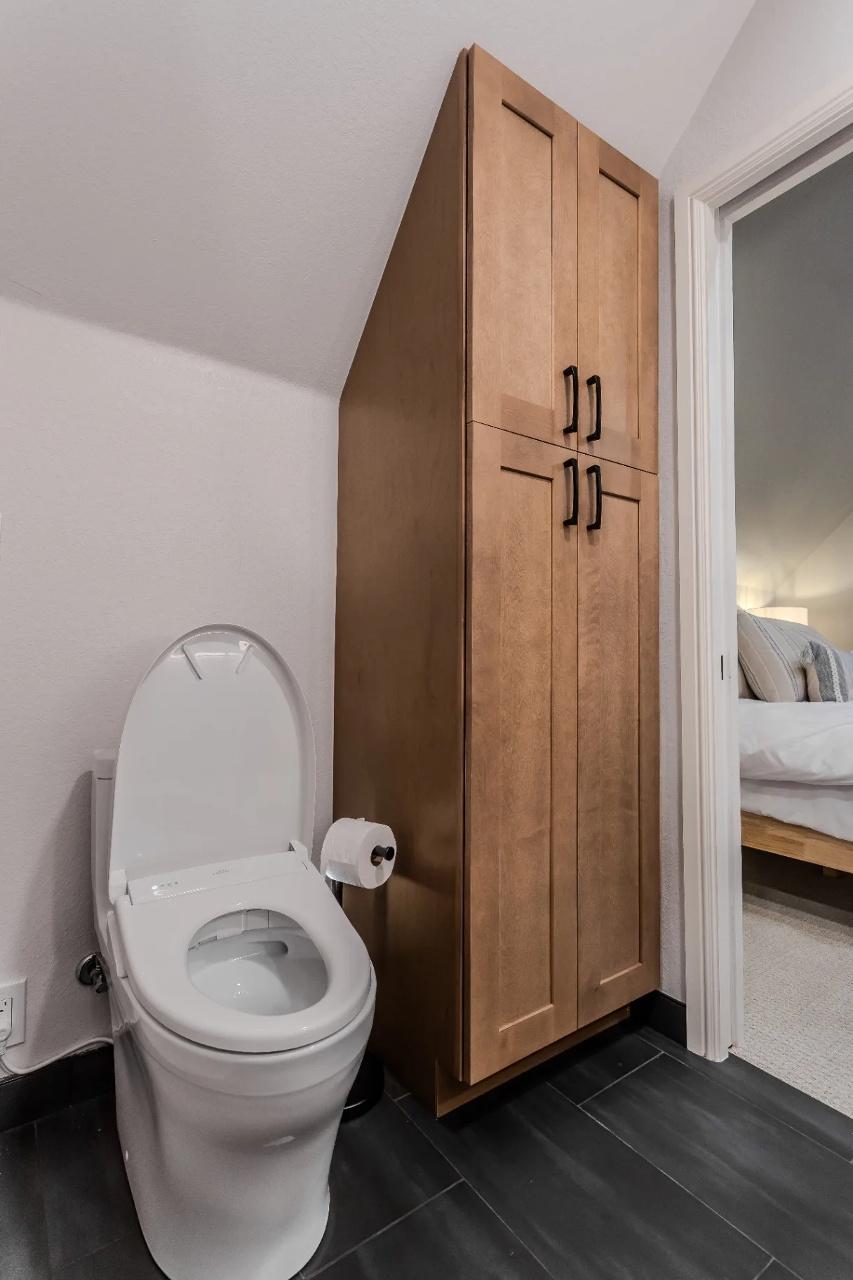 Custom floor-to-ceiling maple linen tower beside a comfort-height toilet on new charcoal tile flooring